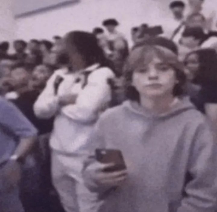 A grainy photo of a young woman in a hoodie holding a smartphone amid a crowded indoor setting.