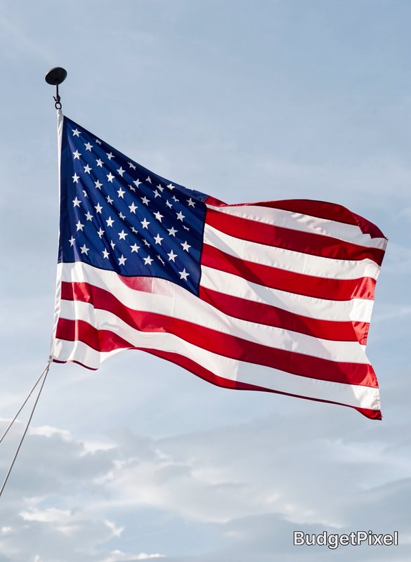 Waving United States American flag mounted on a flagpole, billowing against a soft pale blue clou...