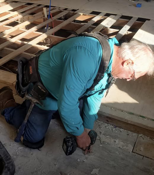 Older man in teal shirt kneeling and drilling on a dusty floor with exposed wooden joists.