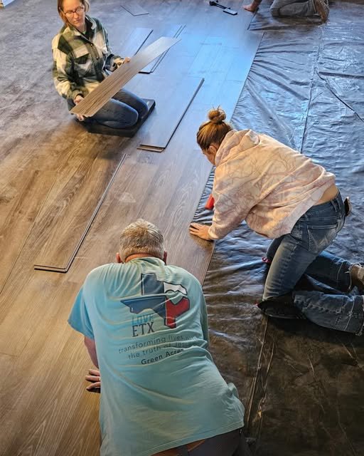 Three people install laminate flooring, aligning planks over black underlayment on a workspace.