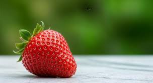 Close up of a ripe red strawberry with green leafy tops resting on a light wood surface against b...