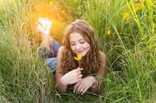 Smiling young woman lies in tall sunlit grass holding a yellow wildflower during golden hour.