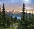 Sunset mountain view framed by tall evergreen pine trees, with an alpine lake and distant rocky s...