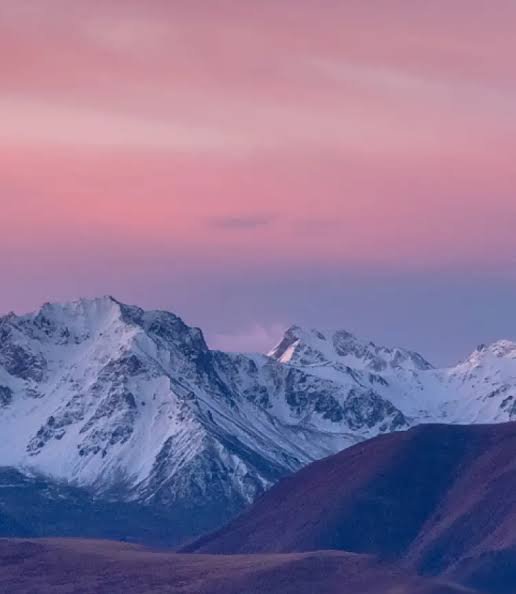 Jagged snow-capped mountain peaks beneath a soft gradient pastel pink and purple sunset sky, roll...