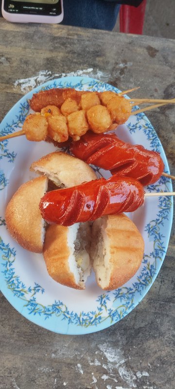 Plate with sausage skewers and fried potato balls next to a split bread bun on a blue floral plate.