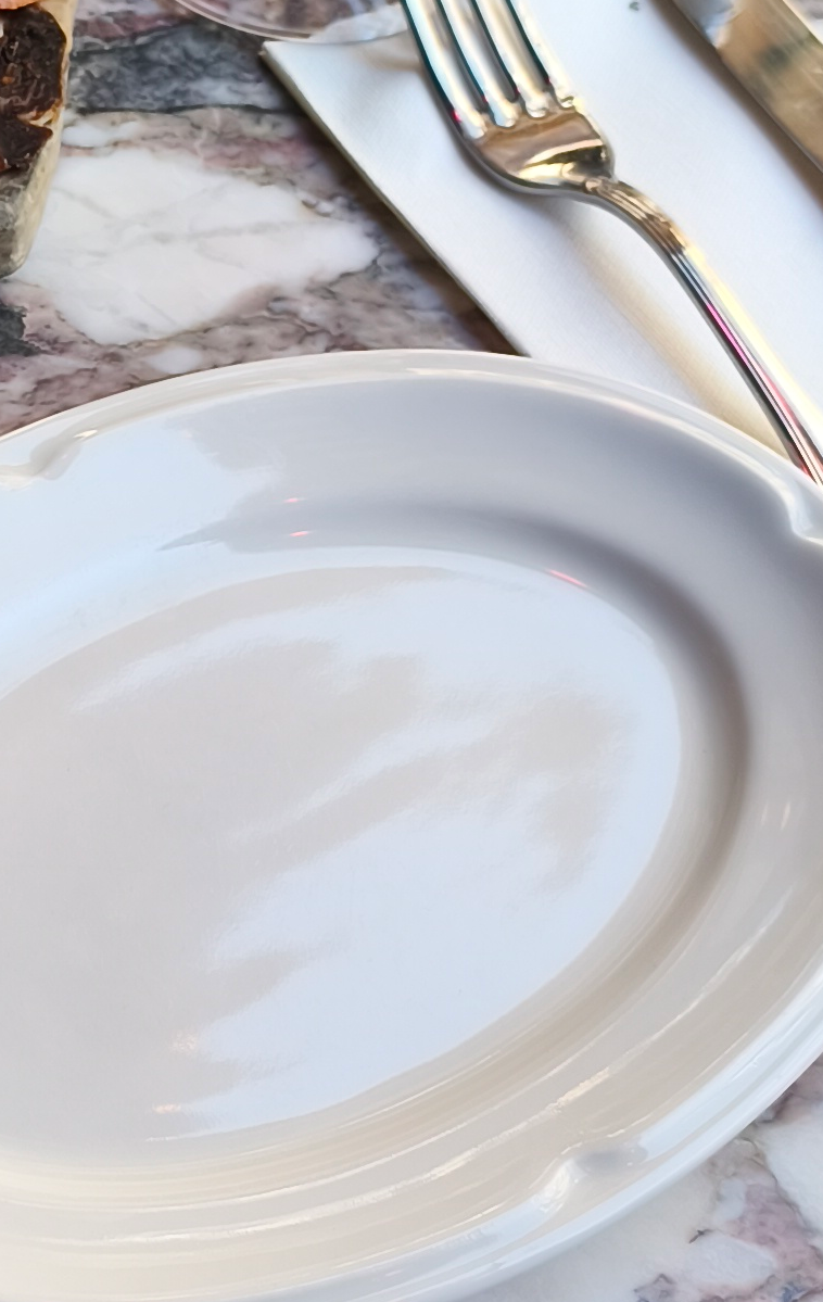 Close-up of an empty glossy white oval dinner plate on a veined marble restaurant table, with a silver fork resting on a w...