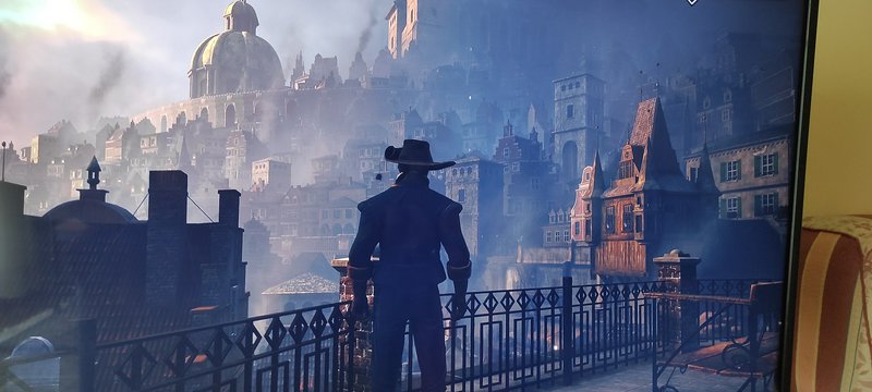 Silhouette of a man in a wide-brim hat on a wrought-iron balcony overlooking a foggy, domed historic city.