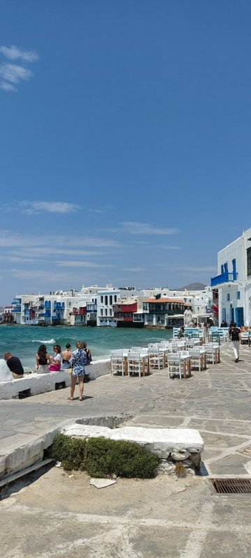 Sunny Mykonos Little Venice waterfront with iconic white Cycladic buildings, tourists, and outdoo...