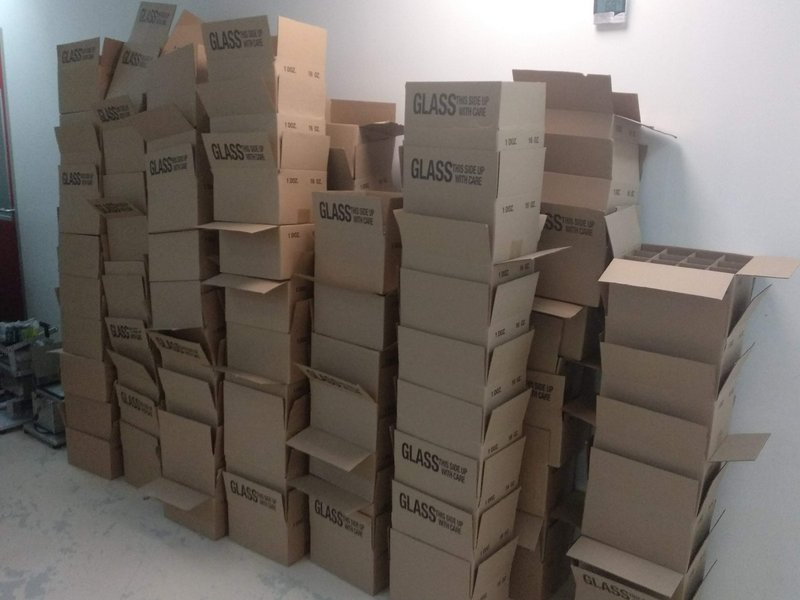 Tall uneven stacks of open cardboard boxes marked "GLASS THIS SIDE UP WITH CARE" piled against a ...