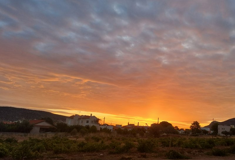 Layered orange and purple sunset sky over a quiet Mediterranean village and rural farm field at golden hour.