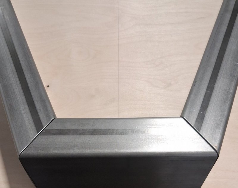 Close-up overhead view of a welded raw steel V-shaped table leg resting on light birch plywood