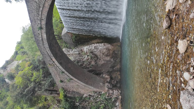 Weathered historic stone arch bridge beside a small waterfall spilling into a clear rocky stream in a lush forested gorge