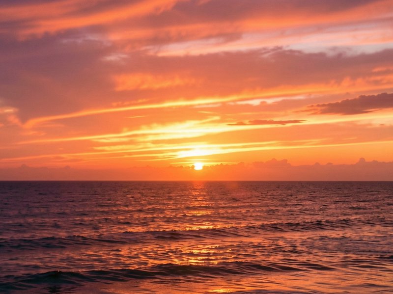 Bright golden sunset glows over rolling ocean waves beneath streaked orange and pink clouds