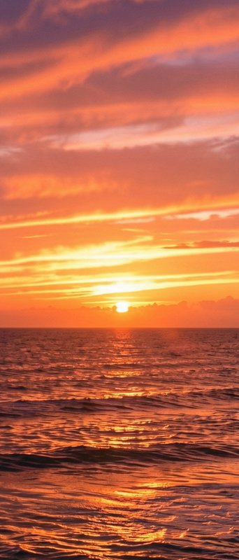 Glowing orange and purple sunset hangs low over rippling open ocean under streaked wispy clouds