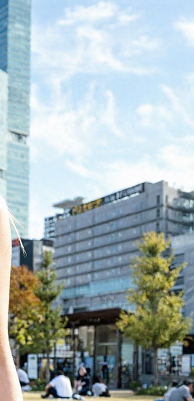 Blurred sunny urban park view, partial foreground arm, modern office buildings, people relaxing o...