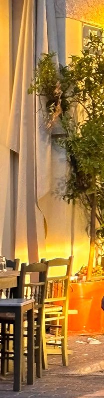 [Warm golden hour sidewalk cafe patio with wooden dining chairs, a potted leafy tree, and draped awning against a plaster ...