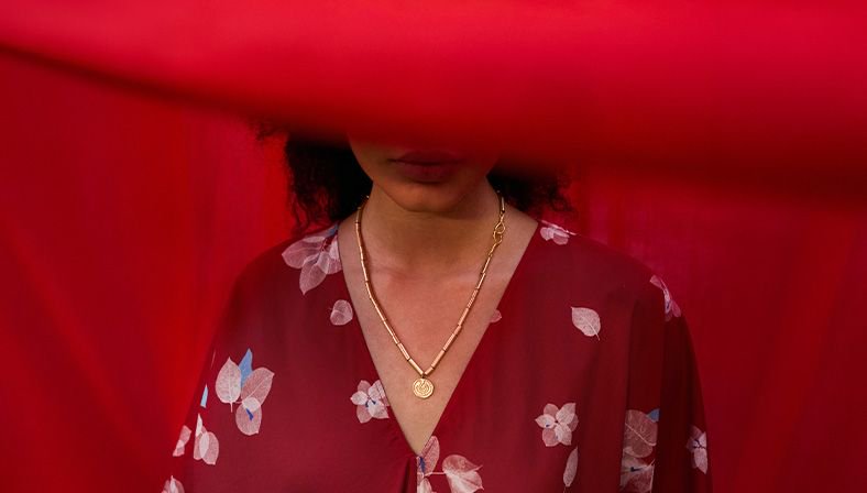 Woman in a burgundy floral dress behind sheer red fabric with a gold pendant necklace