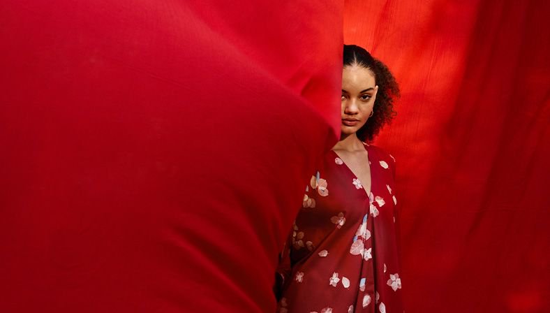 Young woman peeking from behind a red curtain, wearing a red floral dress.
