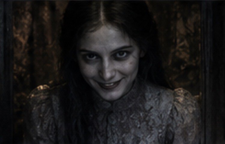 Creepy pale ghostly woman with sunken dark eyes grins menacingly from a dim shadowy old interior