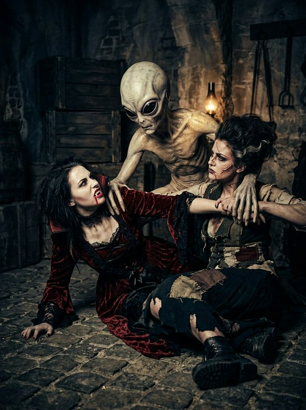 Classic grey alien restrains a fanged vampire woman and an injured companion in a dim cobblestone dungeon