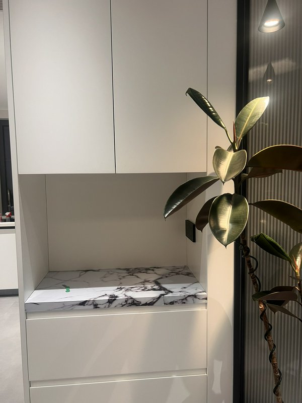 Modern white kitchen cabinet with marble-topped open nook and a tall leafy plant nearby.