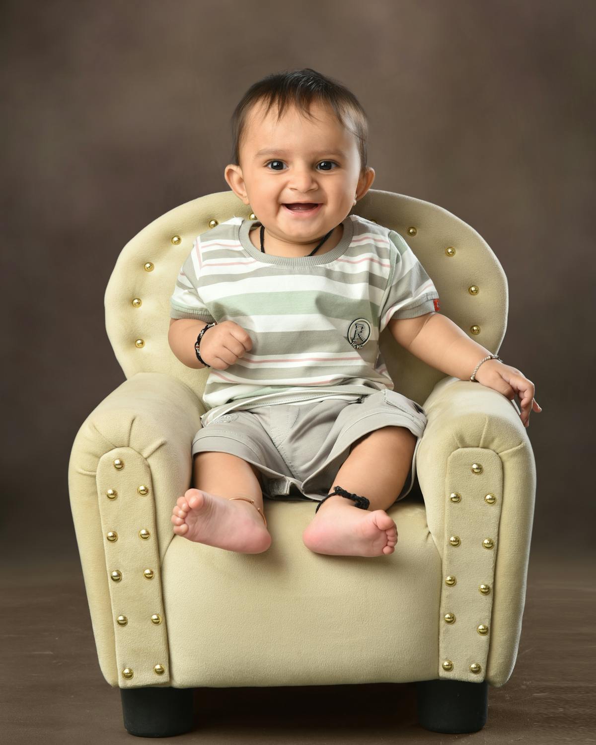 Baby boy smiling in a cream tufted chair with gold studs, wearing striped shirt and shorts.