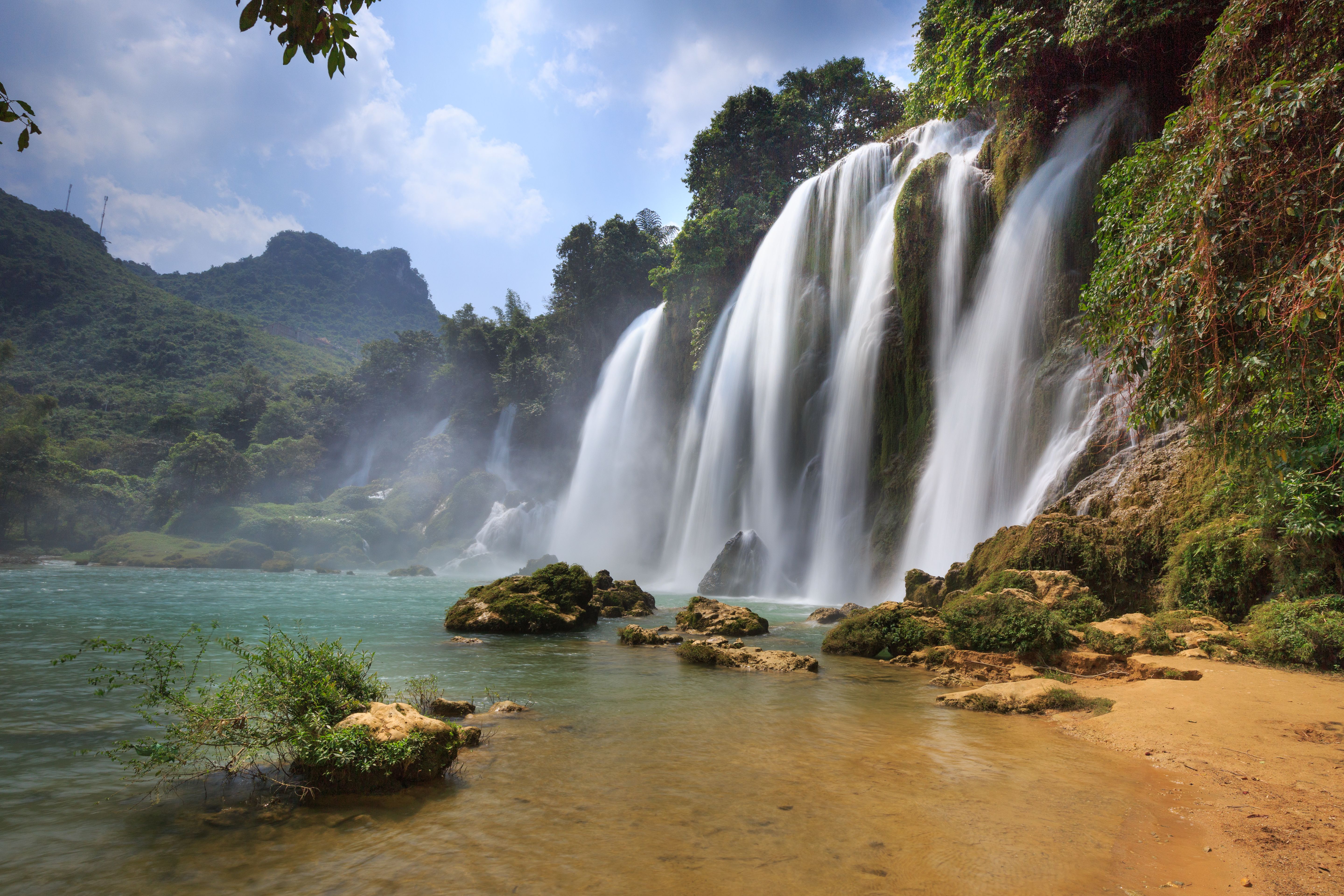 Wide tropical waterfall cascading over rocky cliff into a clear pool with lush jungle and mountains.