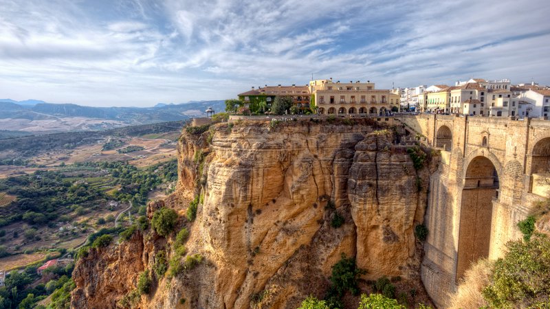 Cliffside town with an arched stone bridge and buildings atop a dramatic rock cliff.