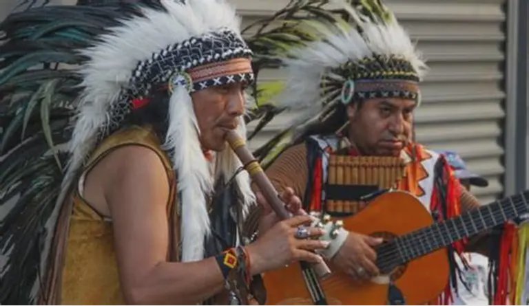 Two Indigenous musicians in feather headdresses playing a flute and a guitar.
