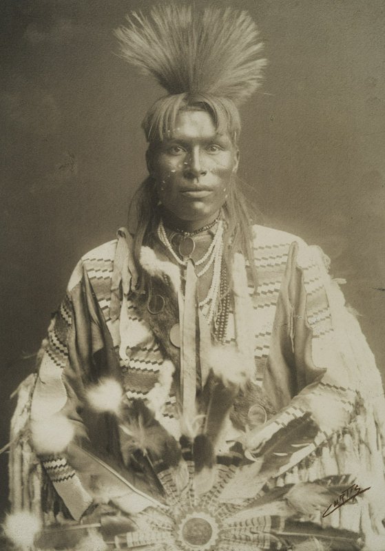 Portrait of a Native American man in ceremonial regalia with a feather headdress.
