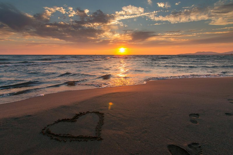 Sunset over the ocean with a heart drawn in wet sand and footprints on the shore.