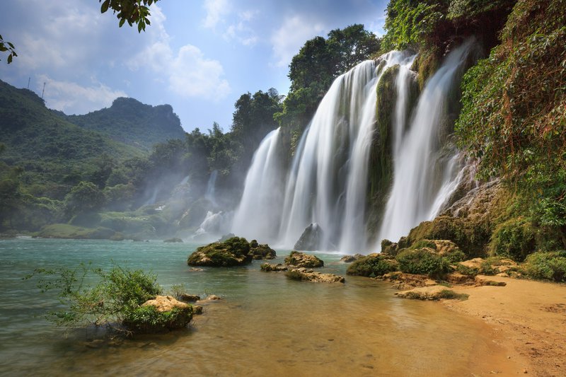 Wide tropical waterfall cascading over rocky cliff into a clear pool with lush jungle and mountains.