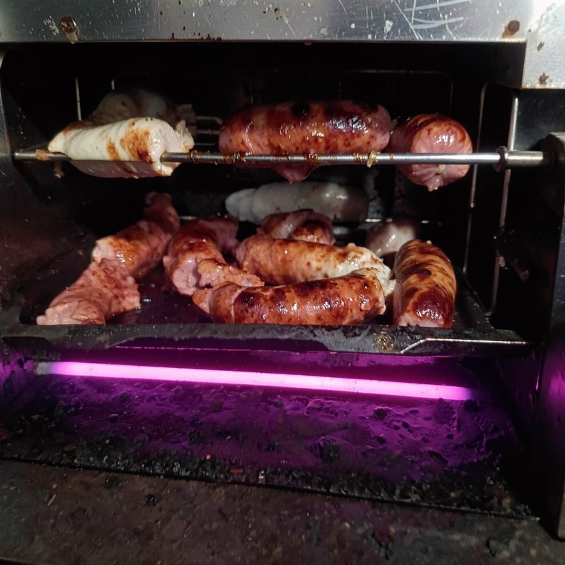 Sausages cook on a rotisserie spit and lower tray inside an electric infrared grill with purple g...