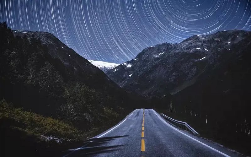Night road through mountains under star trails in a long-exposure sky.