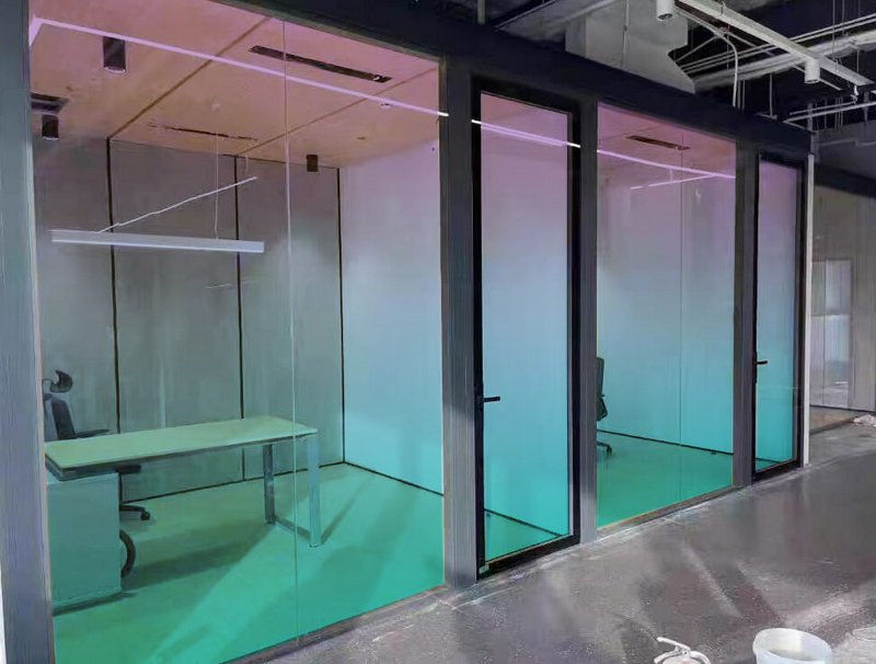 Glass-walled modern office pods with gradient teal lighting and a desk inside.