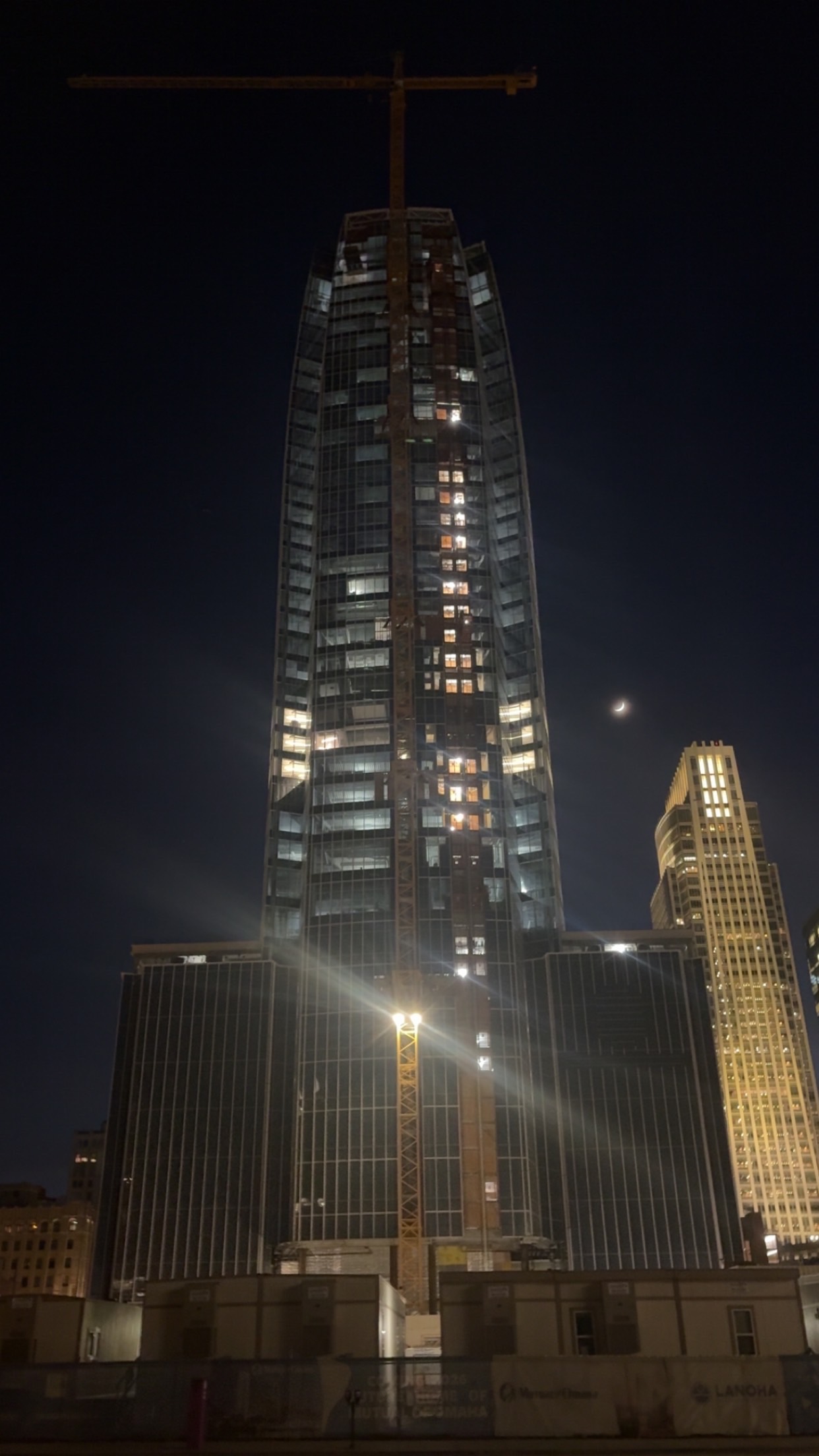 Nighttime construction of a tall glass skyscraper with a crane on top and illuminated windows.