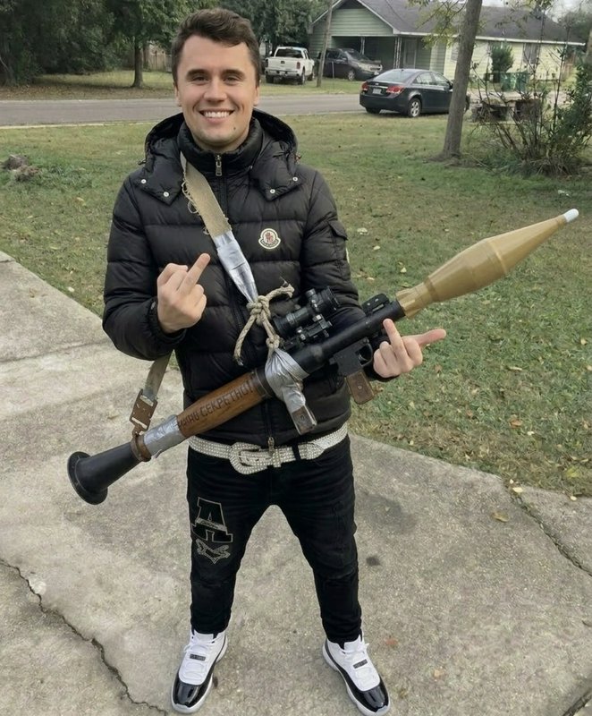 Smiling man in a black puffer jacket outdoors holding a large prop rocket launcher with taped sec...