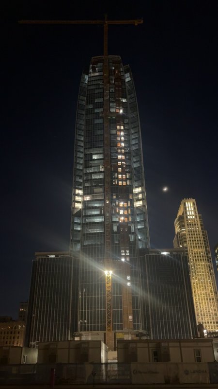 Nighttime construction of a tall glass skyscraper with a crane on top and illuminated windows.