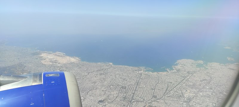 Aerial view from an airplane window showing a blue jet engine, sprawling coastal city, and calm b...