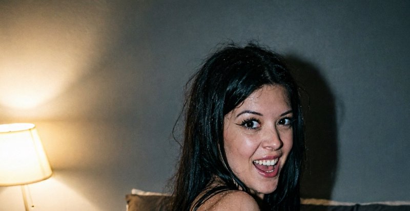 Excited young woman with long dark hair looks over her shoulder, beside a glowing warm bedside la...