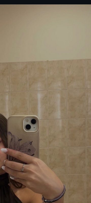 Partial mirror selfie of a hand holding a floral beige iPhone, with white manicured nails, agains...