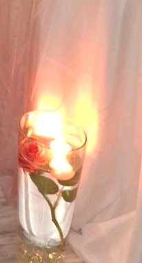 Glowing lit floating candle in a glass vase holding a pink rose, set against soft sheer peach dra...