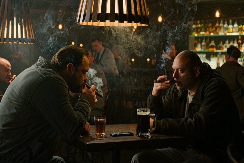Two men smoking cigars sit across a pub table with drinks, in a dim, hazy warm-lit cigar bar.