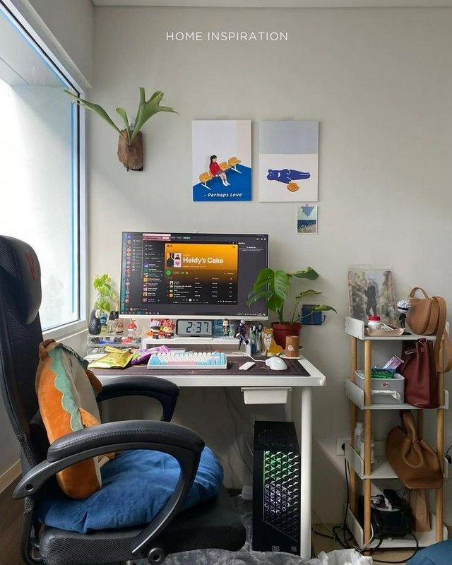 Cozy lived-in home office gaming desk setup with PC, houseplants, wall art, and ergonomic chair n...