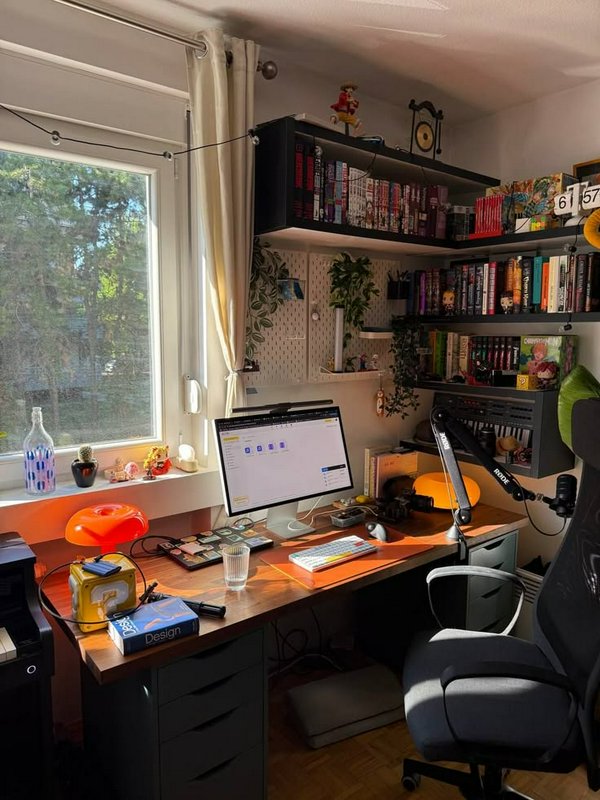 Cozy sunlit home office desk setup with wall-mounted manga shelves, gaming gear, and a window ove...