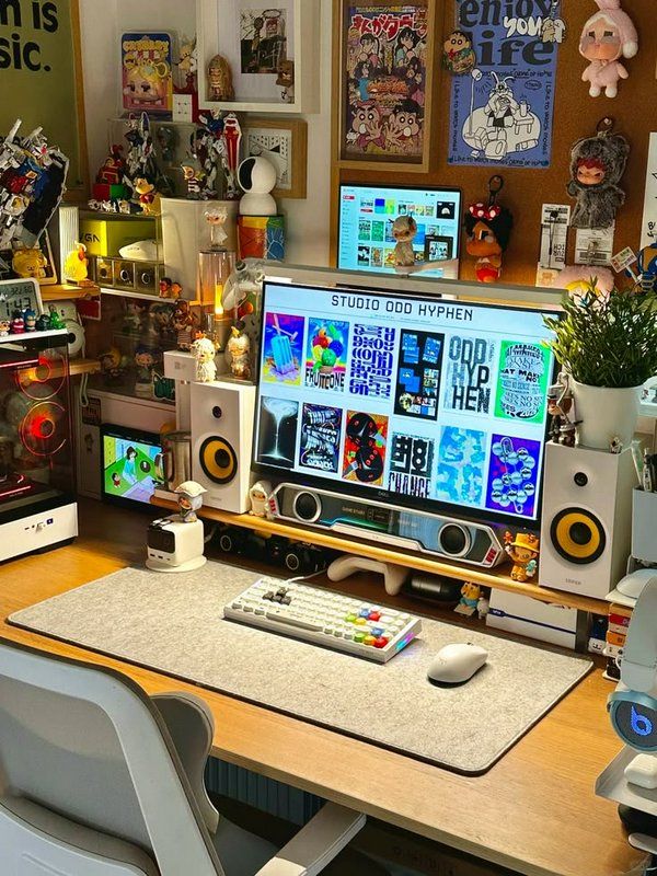 Cozy maximalist gaming desk setup lined with anime figurines, art posters, dual monitors, and a p...