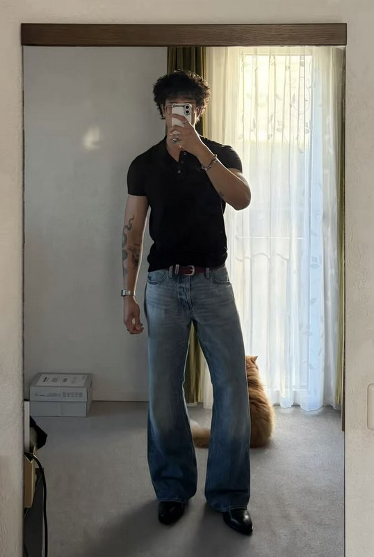 Tattooed curly haired man takes mirror selfie, fluffy orange longhair cat sits behind him near su...