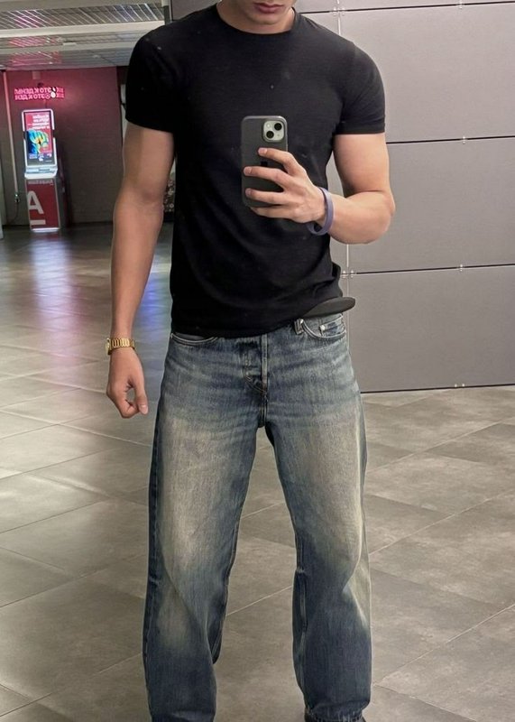 Man takes mirror selfie wearing a fitted black t-shirt, faded wide leg jeans, and casual accessor...