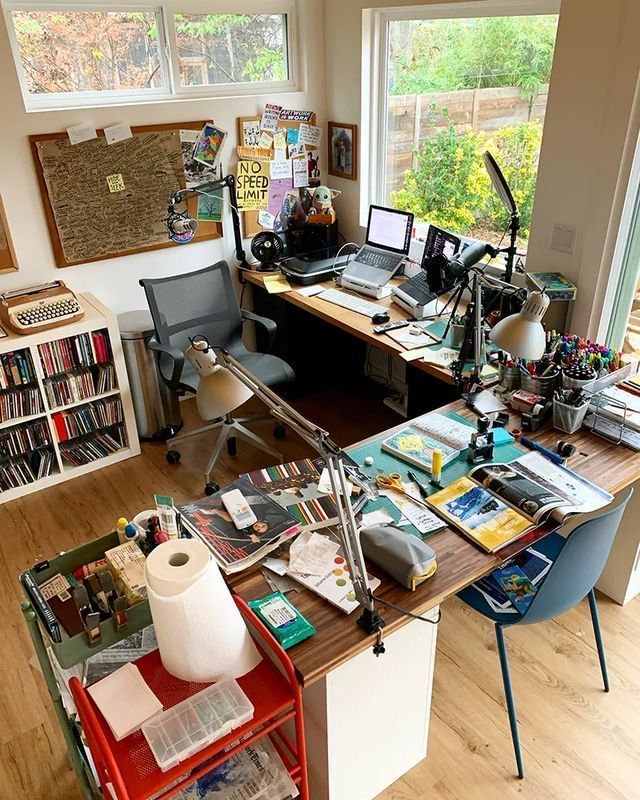 Overhead view of a sunlit, cluttered creative home office workspace with desks, craft supplies, a...