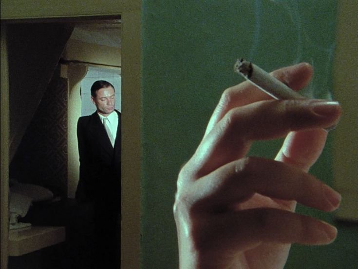 Close-up of a hand holding a lit cigarette, with a suited man standing in a doorway in the backgr...